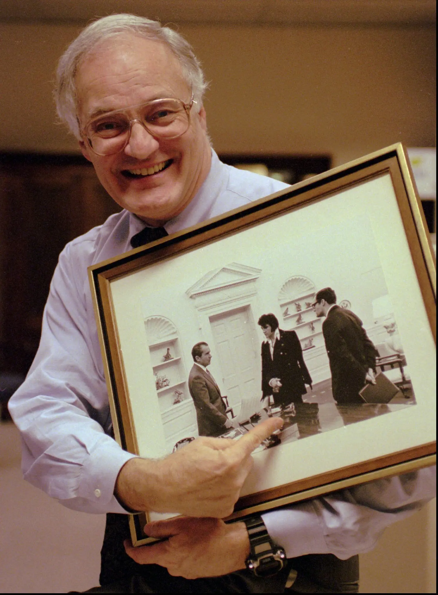 O Sr. Krogh em 1995 em seu escritório em Seattle segurando uma foto emoldurada do encontro de Elvis Presley com o presidente Richard M. Nixon no Salão Oval. O Sr. Krogh, à direita na foto, havia marcado o encontro.Crédito...Associated Press